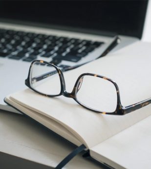 A pair of eyeglasses sits on a desk next to a laptop computer